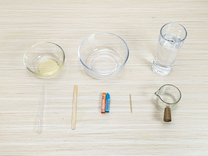 An assortment of items are arranged on a light woodgrain table, seemingly for a science experiment or demonstration. These include: a small glass bowl containing oil, a larger empty glass bowl, a tall glass of water, a clear pipette or dropper, a wooden craft stick, two crayons (one orange an