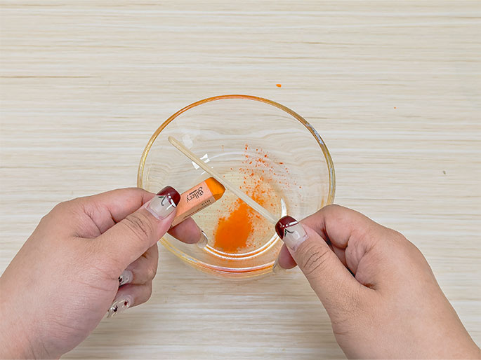 Two hands hold an orange crayon and a wooden craft stick over a clear glass bowl containing a small amount of orange powder or shavings, likely grated from the crayon. The hands appear to be in the process of either grating the crayon with the stick or mixing the crayon shavings into something within the bowl. The bowl sits on a light woodgrain surface. This image likely depicts a craft or science activity.