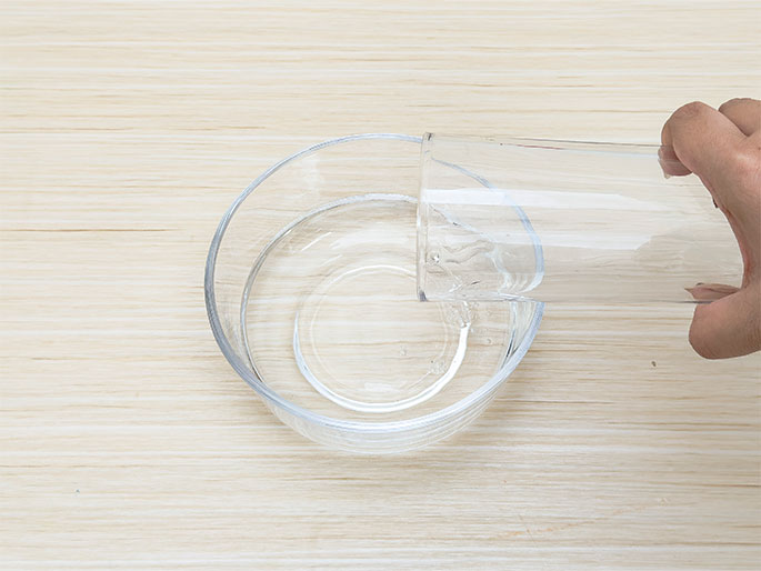 A hand pours water from a tall, clear drinking glass into a clear glass bowl. The bowl already contains a small amount of water. Both the glass and bowl sit on a light woodgrain surface. This image likely depicts a step in preparing for a science experiment, craft, or other activity involving water.
