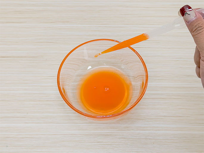 A hand holds a clear plastic pipette containing orange liquid over a small, clear glass bowl. The bowl contains a vibrant orange liquid, and a drop from the pipette is about to fall into it. The bowl rests on a light woodgrain surface. This image likely depicts a science experiment or demonstration involving mixing or measuring colored liquids.