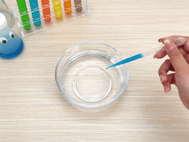 A hand holds a clear pipette containing blue liquid over a clear glass bowl filled with water. The pipette is positioned as if about to dispense the blue liquid into the bowl. In the background, a test tube rack containing test tubes filled with various colored liquids is visible, along with a small, whimsical, googly-eyed bottle or flask. The scene is set on a light woodgrain surface, suggesting a science experiment or demonstration involving color mixing or liquid solutions.