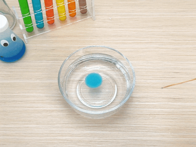 A small, bright blue sphere or blob of a gel-like substance floats in a clear glass bowl filled with water. The bowl sits on a light woodgrain surface. A toothpick is partially visible on the right side of the frame. In the background, a test tube rack with various colored liquids and a small, googly-eyed flask or bottle are visible. This image likely depicts a science experiment or demonstration, possibly illustrating concepts like density or immiscibility.