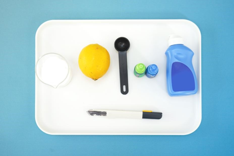 A white rectangular tray sits on a bright blue background, containing the ingredients for a science experiment. The tray holds a lemon, a small glass beaker filled with baking soda, a measuring spoon, two small bottles of food colouring, a bottle of dish soap, a utility knife, and a black marker. The scene is clean and organized, suggesting a controlled and focused environment for scientific exploration.