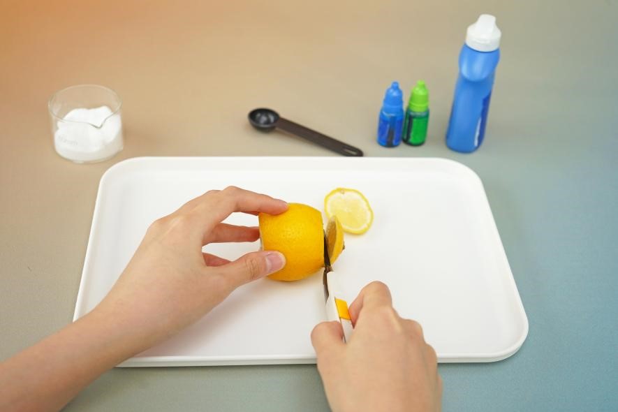 A hand carefully cuts a lemon in half on a white tray. The scene is simple and focused, highlighting the preparation for a science experiment. The lemon, along with the other ingredients visible in the background, suggests a fun and educational activity.