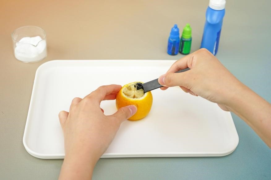 A hand carefully scoops out the inside of a lemon with a black spoon, creating a hollow space. The scene is a close-up, emphasizing the detail of the task and the bright yellow colour of the lemon. The image is a step-by-step guide for a science experiment, capturing the essence of hands-on learning.