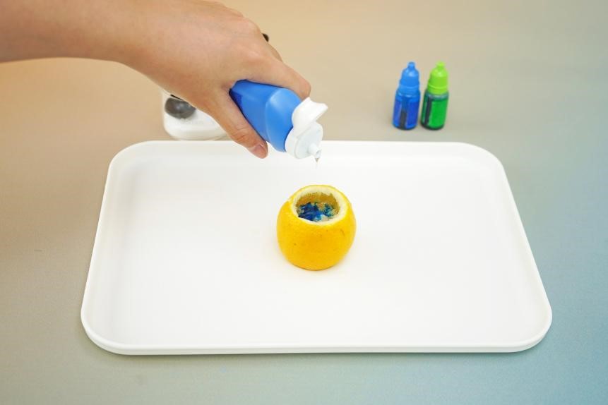 A hand carefully pours dish soap into a hollowed-out lemon, preparing for a science experiment. The scene is simple and focused, highlighting the detail of the task and the bright colors of the ingredients. The image is a step-by-step guide for a science experiment, capturing the essence of hands-on learning.