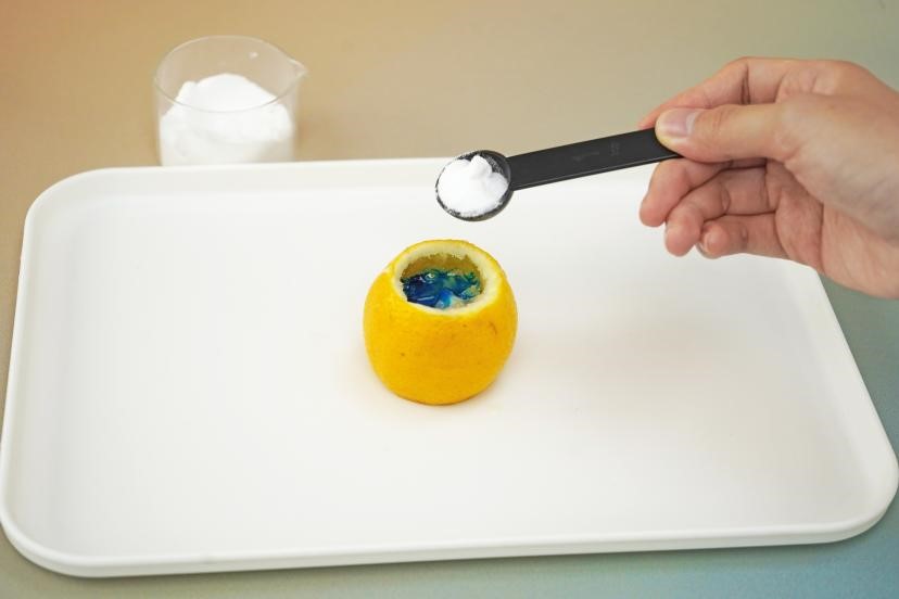 A hand carefully adds baking soda to a hollowed-out lemon with a black spoon, preparing for a science experiment. The scene is simple and focused, highlighting the detail of the task and the bright colours of the ingredients. The image is a step-by-step guide for a science experiment, capturing the essence of hands-on learning.