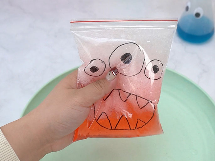A hand holds a sealed, clear plastic zipper bag filled with a bubbly, orange liquid. A simple monster face is drawn on the bag, and the orange liquid and bubbles make the drawing more dynamic. The bag is held above a light green plate, which is partially visible. In the background, a clear container with blue liquid can be seen, likely leftover from a previous step in this craft or science activity. The image suggests the creation of a homemade stress ball, sensory toy, or a simple science experiment involving chemical reactions.