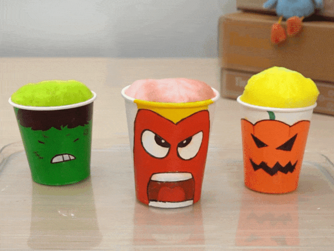Three cups decorated with faces overflow with colorful foam. The first cup is green with a drawing of the Hulk, the second cup is red with an angry face, and the third cup is orange with a jack-o'-lantern face.