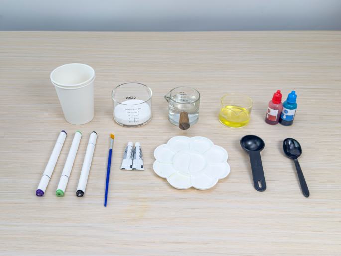 An assortment of art supplies arranged on a light wooden table. The supplies include: a paper cup, three markers, a paintbrush, two tubes of paint, a flower-shaped paint palette, three small glass beakers containing white powder, clear liquid, and yellow liquid respectively, two small bottles of red and blue colouring, a small black measuring spoon, and a regular black spoon. These items appear to be laid out in preparation for an art project.