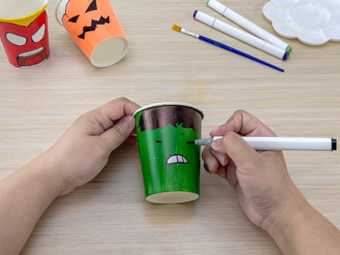 Two hands hold a green paper cup while drawing an angry face on it with a white marker. In the background, other decorated paper cups, markers, a paintbrush, and a paint palette are visible on a wooden table, suggesting an ongoing craft activity. One cup is decorated as a superhero, and another as a jack-o'-lantern.