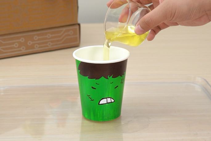 A hand pours a yellow liquid from a clear glass measuring cup into a green paper cup decorated with an angry face. The cup sits on a clear plastic tray, and a cardboard box is visible in the background.