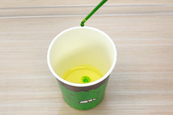 A drop of green liquid from a pipette or dropper is about to fall into a paper cup containing a yellow liquid. The cup is decorated with a drawing of an angry face and rests on a light wooden surface.