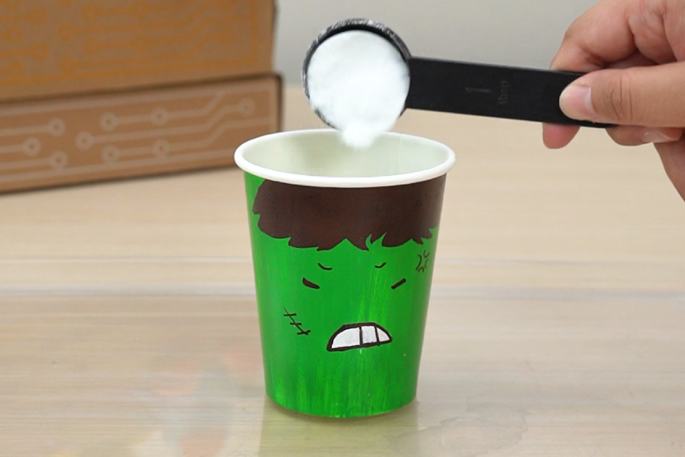 A hand uses a black measuring spoon to pour a white powder into a green paper cup decorated with an angry face. A partially visible cardboard box is in the background on the table. The cup and the action of pouring suggest a science experiment or craft activity is in progress.
