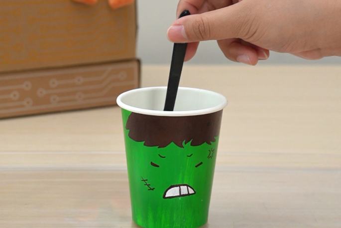 A hand holds a black plastic spoon, stirring the contents of a green paper cup. The cup is decorated with an angry face and sits on a light wooden surface. A partially visible cardboard box is in the background, suggesting this is part of a larger activity or experiment.