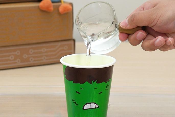 A hand pours water from a clear glass beaker with a wooden handle into a green paper cup decorated with an angry face. A cardboard box is visible in the background on the table. This image likely depicts a step in a science experiment or a similar activity.