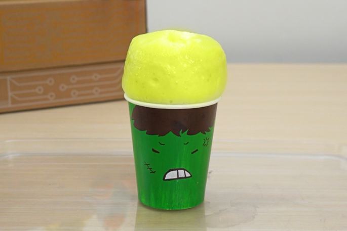 A green paper cup, decorated with an angry face, overflows with a voluminous, light yellow foam. The cup sits on a clear plastic tray, and a cardboard box is visible in the background. This image suggests a science experiment that has created a foamy reaction.