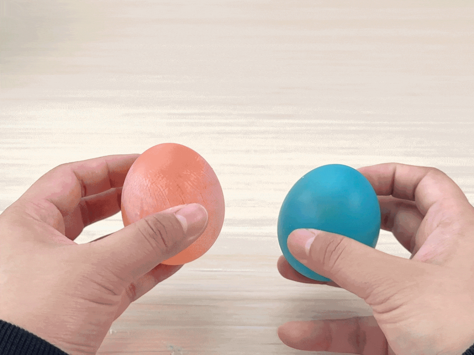 Two hands hold a small, peach-colored ball and a teal-colored ball. The hands gently squeeze both balls, demonstrating their squishy or flexible texture. The balls appear to be made of a soft, rubbery, or gel-like material. The teal ball is then released and bounces slightly on a light wooden surface. The video showcases the tactile properties of two colorful, bouncy balls.