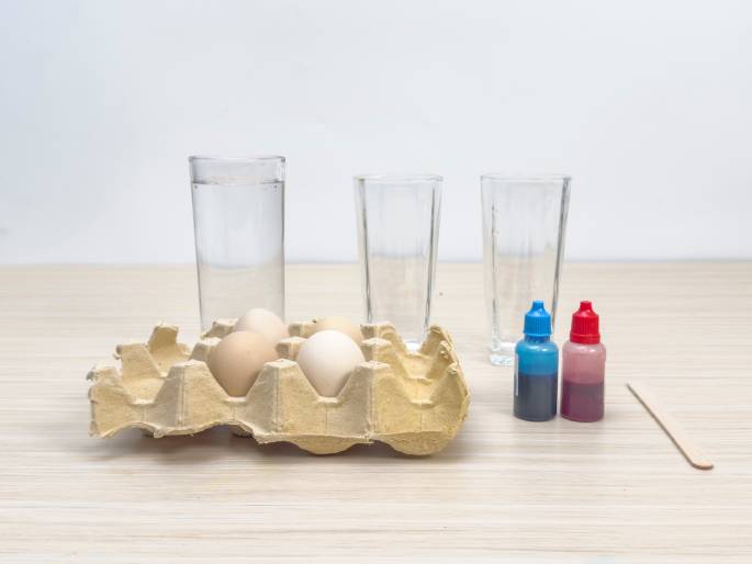 Arranged on a light woodgrain table are items for an activity, likely egg dyeing. A cardboard egg carton holds a few white and light brown eggs. A clear glass filled with water sits behind the egg carton, along with two empty clear glasses. Two small bottles containing blue and red liquid dye are positioned to the right of the glasses. A small wooden craft stick lies flat beside the dye bottles, probably for stirring. The setup suggests preparations for an experiment or craft involving coloring the eggs.