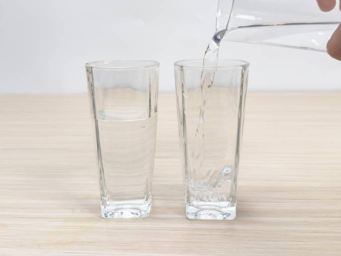 Water is being poured from a clear glass beaker into a clear glass on a light woodgrain table. Another glass with water already in it sits next to the glass being filled. The focus is on the action of pouring the water.