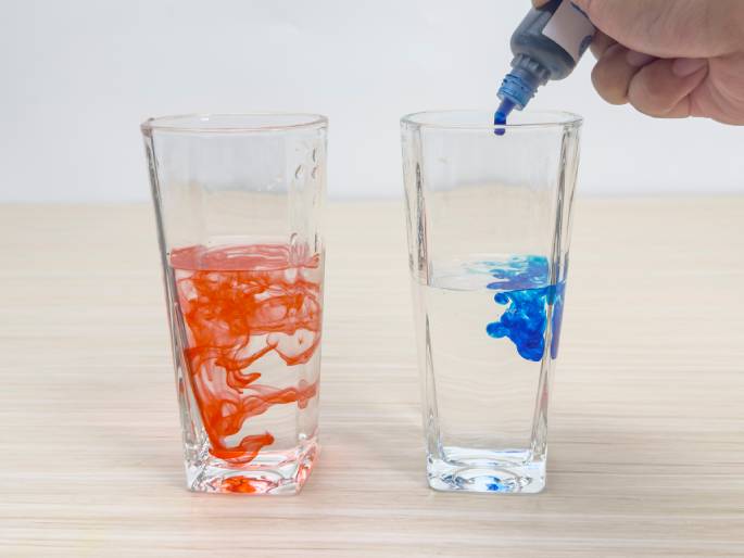 On a light woodgrain surface, two clear glasses contain water. The glass on the left has red dye swirling in it, while a hand is adding blue dye from a small bottle to the glass on the right. The blue dye is visible as a concentrated blob, not yet fully dispersed in the water. This image likely illustrates a color mixing experiment or the process of preparing dye for another purpose.