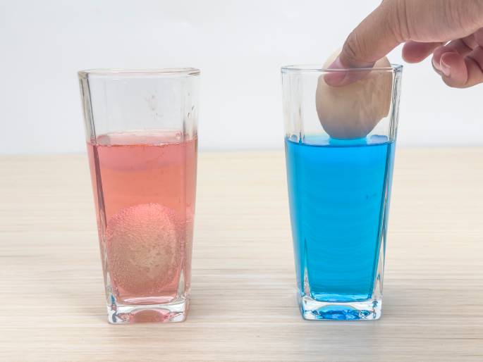 Two clear glasses sit on a light woodgrain table. The glass on the left contains a pink liquid and a submerged egg. The glass on the right holds bright blue liquid, and a hand is placing another egg into the blue dye. This image captures the process of dyeing Easter eggs.