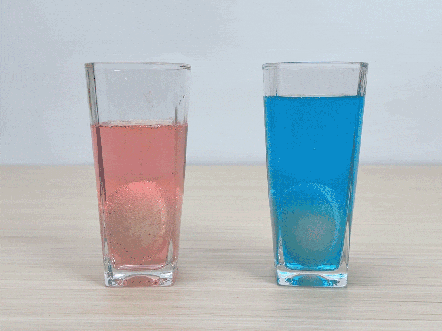 Two clear glasses containing colored water and eggs sit on a light wooden table. One glass holds pink liquid and an egg, while the other contains blue liquid and an egg. Small bubbles cling to the surfaces of both eggs, suggesting a recent immersion or a reaction taking place. This image likely depicts an experiment or demonstration, possibly related to egg dyeing or density.