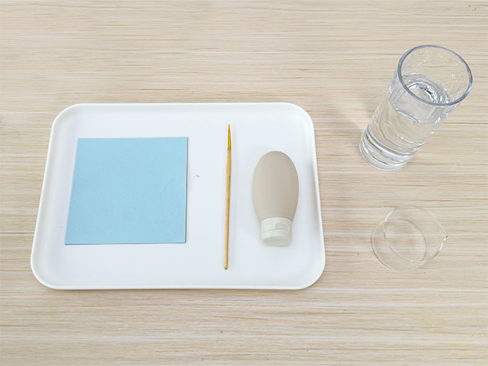 Arranged on a light woodgrain table are art or craft supplies. A white rectangular tray holds a light blue square of paper or card, a small paintbrush or thin wooden stick, and a small beige squeeze bottle or tube. Beside the tray, a clear glass filled with water and an empty, clear, shorter cylindrical container, possibly another glass or jar, are visible. This image likely depicts the setup for a painting, drawing, or other craft activity.