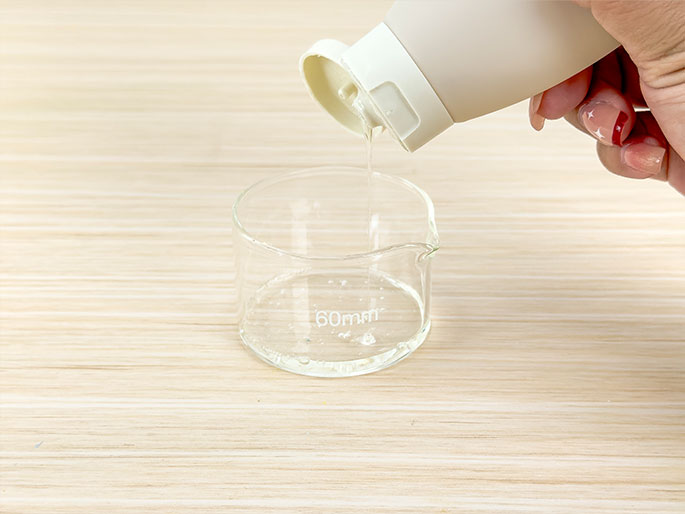 A hand pours a clear, viscous liquid from a beige, plastic squeeze bottle with a white flip-top cap into a small, clear glass beaker or measuring cup. The beaker sits on a light woodgrain surface and has a 60mm marking, likely indicating its diameter. A small amount of liquid is already present in the bottom of the beaker. This image likely depicts a step in a science experiment, craft project, or other activity requiring precise measurement of liquids.