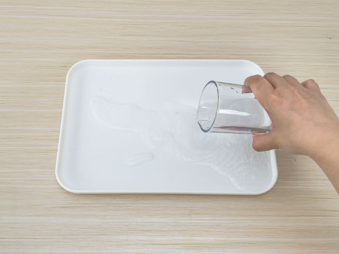 A hand pours water from a clear drinking glass onto a white rectangular tray. The water is spreading across the tray's surface. The tray rests on a light woodgrain table or countertop. This image could be depicting part of a science experiment, a cleaning process, or an art project.