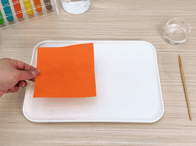A hand holds a bright orange piece of paper over a white tray. The word Tinkerer is faintly visible, written in a darker orange on the paper. The tray sits on a light woodgrain surface. A small paintbrush and a clear glass or jar of water are visible beside the tray. This scene appears to be part of an art or craft activity. A test tube rack with colorful liquids is visible in the background, suggesting a science experiment or demonstration as well.