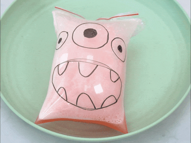 A clear plastic zip-top bag with a cartoon monster face drawn on it rests in a shallow, light green bowl. The bag contains a pink, bubbly mixture that quickly expands, overflowing the bag and spilling into the bowl. The mixture changes color from pink to light blue as it expands, creating a foamy overflow. This suggests a chemical reaction is taking place within the bag.