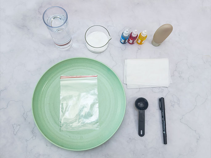 An assortment of materials is laid out on a marbled surface, suggesting preparation for a science experiment or craft project. These include a glass of water, a small beaker containing a white powder, three small bottles of primary color dyes (blue, red, and yellow), a beige squeeze tube, a light green plate with a small, clear zippered plastic bag containing a white powder, a few folded white paper towels or tissues, a small black measuring spoon, and a black marker.