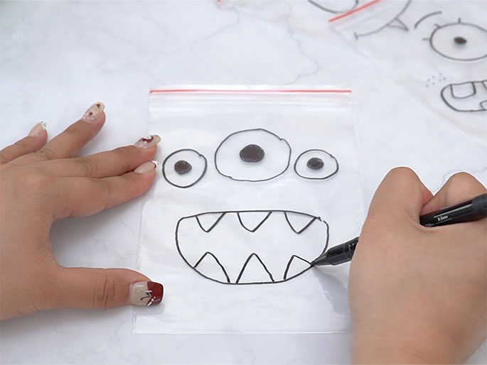 Two hands are engaged in creating a monster drawing on a clear plastic zippered bag. One hand holds the bag while the other draws triangular teeth inside a wide, open mouth with a black marker. Three eyes, each circled in black with a smaller black dot in the center, are already drawn above the mouth. Other similar bags with different monster faces are partially visible in the background, suggesting a craft activity involving multiple monster creations.