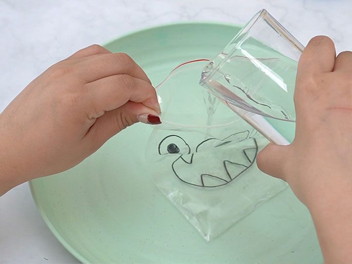 Two hands are depicted in the process of a science experiment or craft activity. One hand holds open a clear plastic zipper bag with a simple monster face drawn on it, while the other hand pours water from a clear glass into the bag. The bag is positioned over a light green plate or shallow bowl. The monster face consists of one eye and a toothy grin. This image likely illustrates a step in creating a sensory toy or a simple science demonstration.
