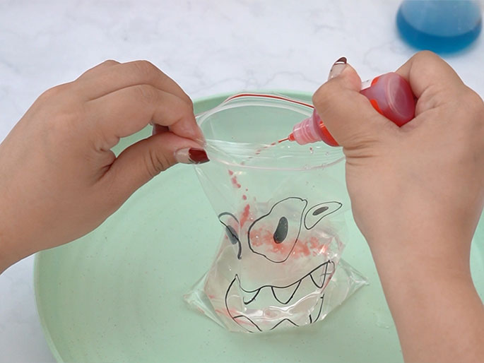 Two hands are shown adding red coloring to a clear plastic bag containing a clear liquid. The bag, which has a monster face drawn on it, rests on a light green plate. The monster face now has red coloring inside the outlines of its eyes, creating a more defined and vibrant image. A small bottle with a nozzle, presumably containing the red dye or paint, is held by one hand, squeezing drops of color into the bag. This continues the process of creating a sensory bag or a science experiment demonstrated in the previous images. A bottle of blue liquid is visible in the background, likely to be added next.