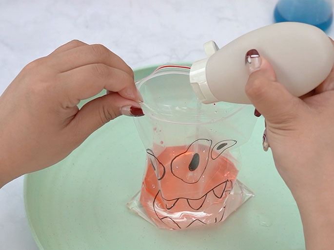 Two hands are interacting with a clear plastic zipper bag containing a pinkish-orange liquid. The bag, which has a monster face drawn on it, rests on a light green plate. One hand holds the bag open while the other squeezes a light-colored, opaque, squeezable bottle, adding more liquid to the bag. The monster's eyes are now filled with the pinkish-orange color. This image suggests a continuation of the process shown in previous images, likely the creation of a sensory bag or a similar craft/science project. A blue liquid is visible in a container in the background, hinting at the next step.