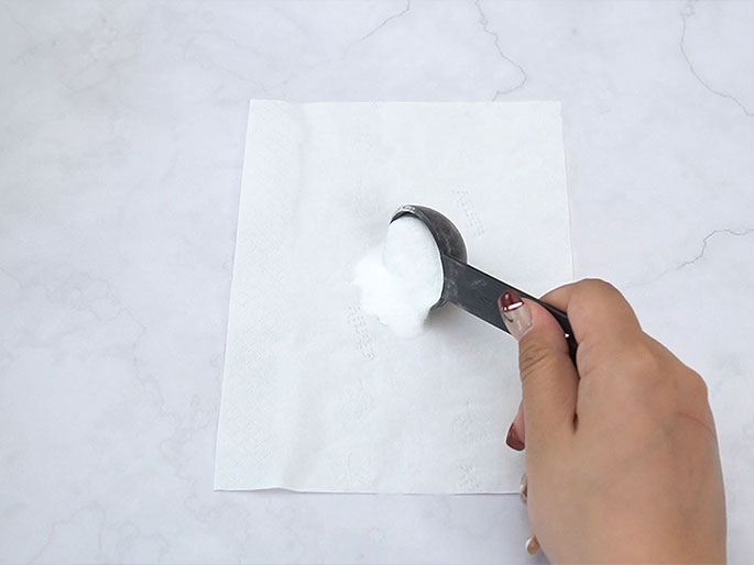 A hand holds a black measuring spoon filled with a white powder, likely baking soda or a similar substance, over a white paper towel. Some of the powder has already spilled onto the towel. The background is a marbled white surface. The image suggests a preparation step in a recipe, experiment, or cleaning activity.