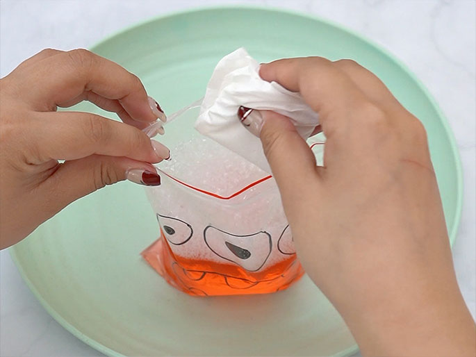Two hands are adding a folded white paper towel or tissue into a clear plastic zipper bag. The bag, which sits on a light green plate, contains a bubbly orange liquid and is decorated with a monster face. One hand holds the bag open while the other inserts the paper towel. This image seems to be part of a sequence demonstrating the creation of a sensory toy or a science experiment, where the wrapped powder from the previous images is now being combined with the liquid.