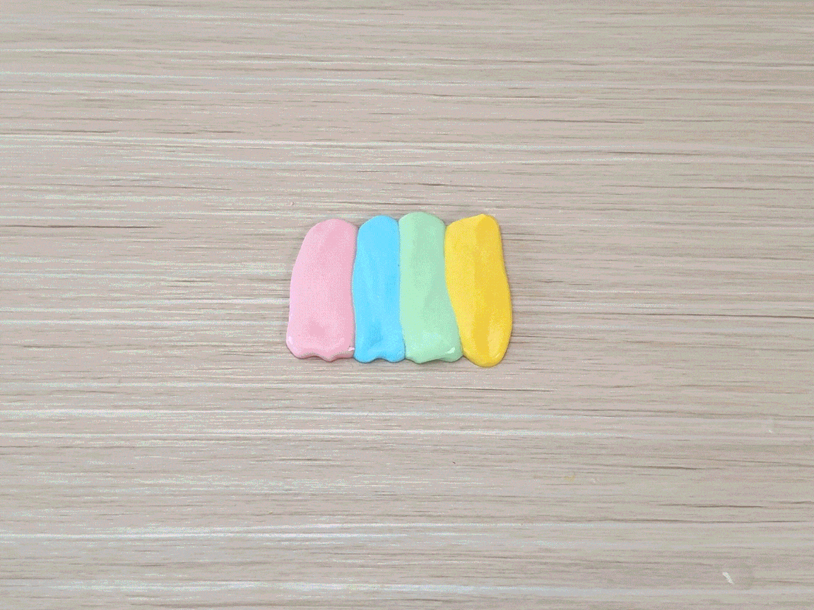 Two hands press down on four parallel lines of pastel-colored slime or playdough on a light wooden surface. The colors are pink, light purple, light green, and yellow. The hands then gather the slime/playdough together and stretch it, revealing the swirling pastel colors inside. The video demonstrates the playful manipulation of a colorful, stretchy substance, likely slime or a similar material.