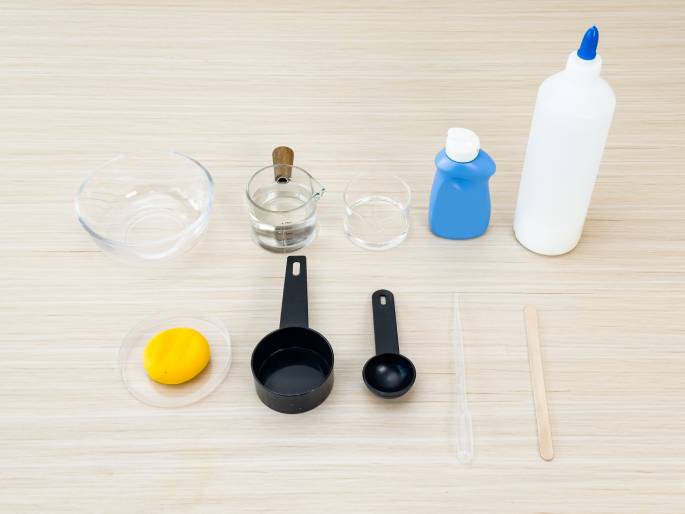An assortment of items are arranged on a light woodgrain table, suggesting preparation for a science experiment or craft activity. The items include: a clear glass bowl, a small glass measuring cup with water, an empty short clear glass, a small blue plastic bottle, a taller white plastic bottle with a nozzle tip, a small clear dish with a yellow substance (possibly playdough or slime), a larger and smaller black measuring spoon, a clear pipette or dropper, and a wooden craft stick.