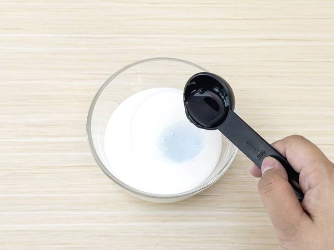 A hand holds a black measuring spoon containing a clear liquid over a clear glass bowl filled with a white substance, likely glue. A small amount of the clear liquid has already been added to the bowl, as evidenced by a slightly translucent area in the white substance. The bowl sits on a light woodgrain table. This image appears to depict a step in making slime or another craft project.
