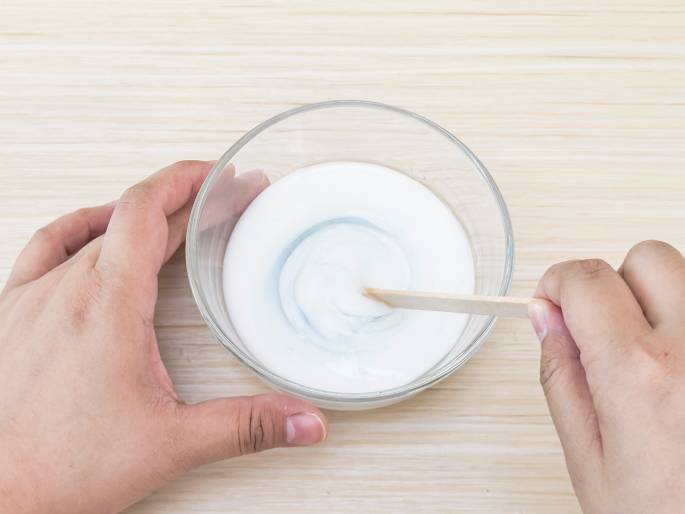 Two hands interact with a clear glass bowl containing a white substance, likely glue, on a light woodgrain table. One hand steadies the bowl while the other stirs the mixture with a wooden craft stick. A swirling pattern is visible in the glue, indicating the mixing process. A slight bluish tint within the glue suggests that another liquid has been added. This image likely depicts a step in a craft project, such as making slime.