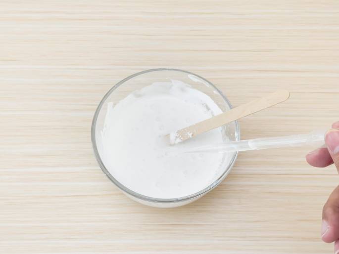 A clear glass bowl containing a thick, white mixture sits on a light woodgrain table. A wooden craft stick rests partially submerged in the mixture, and a hand holds a clear pipette or dropper above the bowl, likely having just added or about to add more liquid. The mixture has a slightly uneven texture, suggesting it is still being combined. This image likely represents a step in a craft or science experiment.