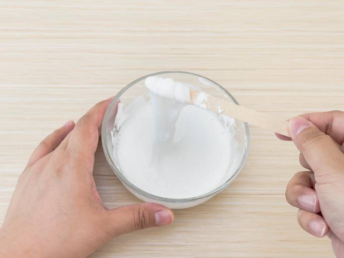 Two hands are shown interacting with a clear glass bowl containing a thick, white, glue-like substance on a light woodgrain table. One hand holds the bowl steady while the other lifts a wooden craft stick out of the mixture. A thick strand of the white substance stretches from the stick back down to the bowl, demonstrating its viscous nature. This image likely illustrates a stage in making slime or a similar craft project.