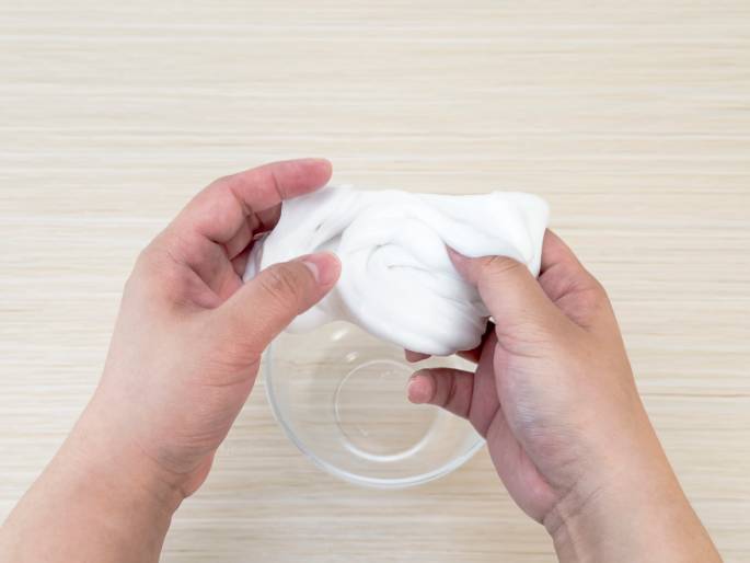 Two hands hold a mass of white slime above an empty clear glass bowl on a light woodgrain table. The slime is being gathered and folded, showing its pliable texture. The bowl likely recently held the ingredients or the slime itself during its creation. This image suggests the successful completion of a slime-making activity.