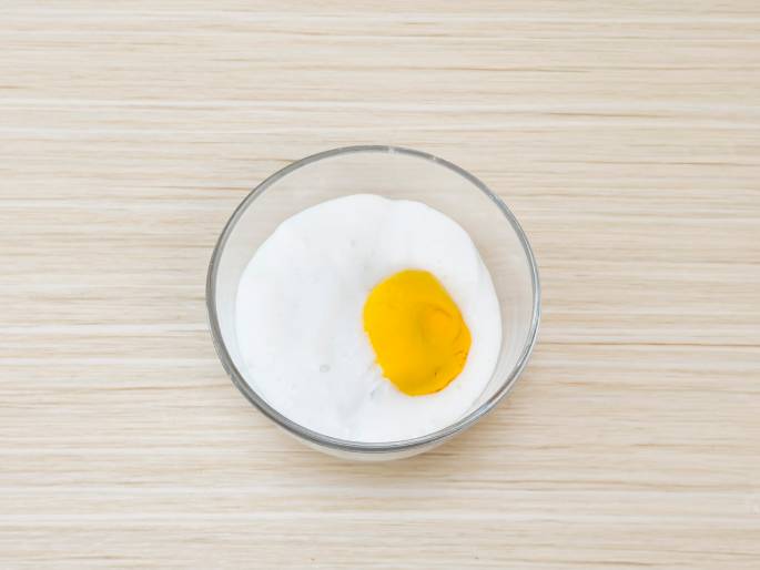 A clear glass bowl sits on a light woodgrain table. Inside the bowl, a white powdery substance, likely baking soda or a similar ingredient, surrounds a small pool of bright yellow liquid. This image likely depicts the initial stages of mixing ingredients for a recipe or experiment.