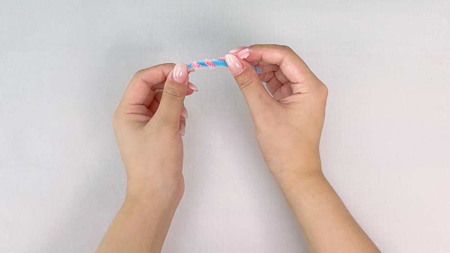 Ooh, look at that! A pair of hands gently twists together two pipe cleaners, one pink and one blue, creating a lovely swirly effect. It's all rather neat, innit? A perfect candy cane in the making!