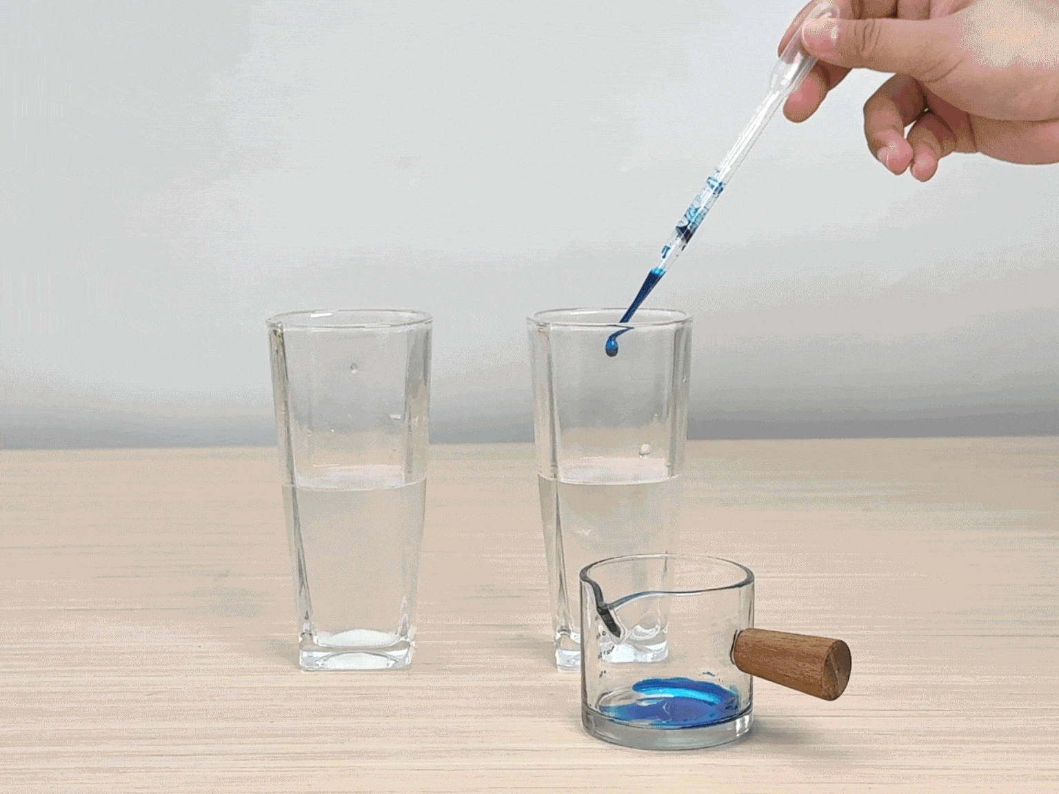 A hand uses a pipette to drop blue food coloring into two clear glasses of water. A small glass pitcher with a wooden handle, also containing blue liquid, sits next to the glasses on a light wooden table. The food coloring disperses differently in each glass; in one, it forms a distinct layer at the bottom, while in the other, it swirls and mixes more readily. The video likely demonstrates a simple science experiment illustrating concepts like density or diffusion.