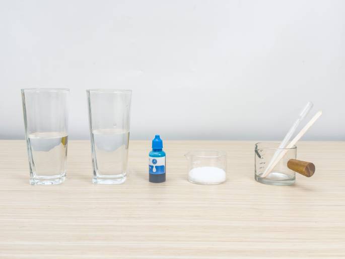 An array of items commonly used in science experiments is arranged on a light woodgrain table against a plain white background. From left to right, there are two clear glasses partially filled with water, a small bottle of blue food coloring, a small glass beaker containing a white powder (possibly baking soda or borax), a clear glass measuring cup with a wooden handle containing a wooden stirring stick and a clear pipette or dropper. This image likely depicts the ingredients and tools gathered before starting a science experiment, possibly making slime.